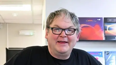 ALEX STEVANOVIC/BBC Damian is in a room with pictures behind him on a wall.
He is wearing glasses.