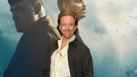 Getty Images Damian Lewis looks at the camera and smiles. He is standing in front of a large picture of two men, in a still from the film Pressure, at its New York Premiere.