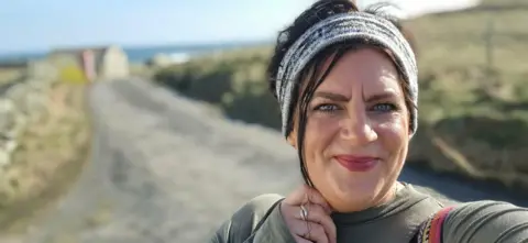 Tammy Hine Tammy standing in a road looking at the camera, with a band round her head. Her hair is tied back. She is wearing a green top, a road and fields are behind her, but blurred. 