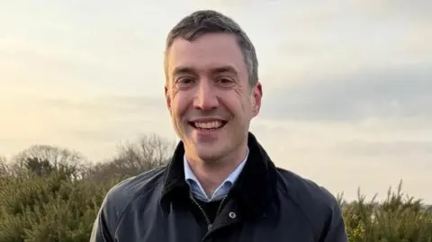 Nadira Tudor/BBC Adrian Ramsey smiling at the camera against the background of some shrubbery. He is wearing a grey waterproof coat and has dark hair.