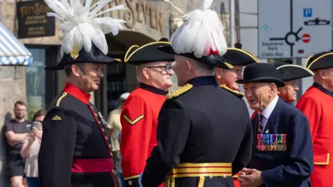 BBC People stood together wearing black and red uniforms. All of the people are wearing black hats with yellow rims, and there is two people with feathers