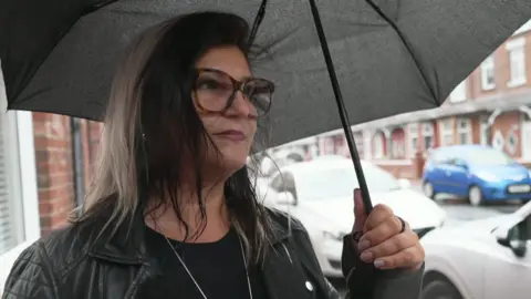 Lili Myers stands in a residential street wearing a black leather jacket and she holds an umbrella. It is raining hard. She has brown hair and is wearing tortoise shell glasses.