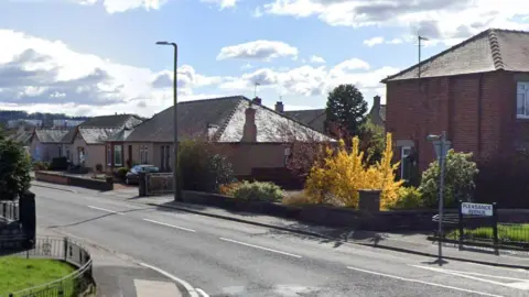 Google A town street with a sign in the foreground and two brown brick houses with trees cars in the driveways and a blue cloudy sky on a sunny day