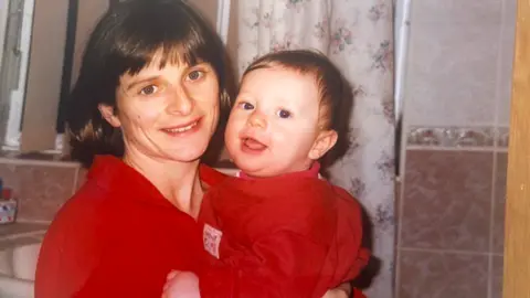 Family Handout A woman is holding her baby son in her arms. Both mother and son are dressed in red and smiling at the camera. The woman's hair is brown and cut in a bob, with a fringe, and pink bathroom tiles and a floral shower curtain can be seen behind.