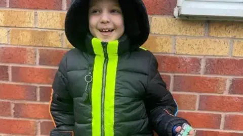 Charlie smiles at the camera. He is wearing the Chammo coat which is dark green and padded and there is a large hood over Charlie's head. The coat has a large high visibility stripe along the zip.
