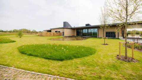 One storey building made of beige brick with large dark windows. There is grass in front with wildflowers, two trees and a path. There is also a pond and then trees in the distance.