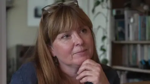 A woman with long strawberry-blonde hair and fringe looks thoughtfully away from the camera with her hand on her chin