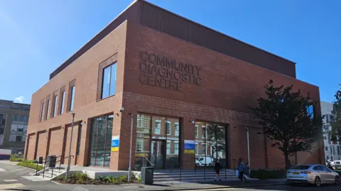 Hull University Teaching Hospitals Hull's Community Diagnostic Centre on Albion Street. A large, modern brick building stands on a street corner and is partially shaded from the sun. Three people are walking past the large glazed entrance which has four steps leading up to the door. A blue sky is visible in the background.