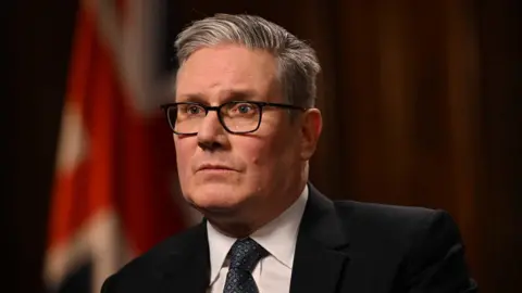 Sir Keir Starmer with a serious expression pictured from the shoulder up, wearing a tie and suit, with neat black rimmed glasses, with a union flag out of focus behind him 
