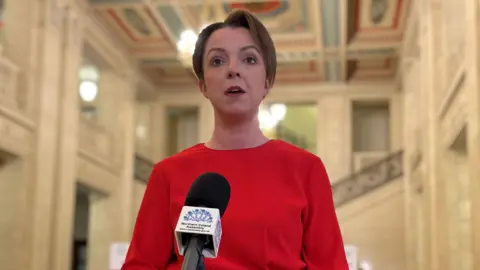 Kimmins is standing in front of a mic in the hall at Stormont. She's wearing red. Marble steps are behind her.