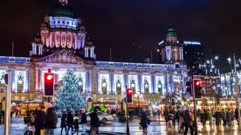 Getty Images Belfast City Hall lit up with bright white stars in every alcove. Stalls from a Christmas market are visible in front, and there is a big Christmas tree lit up with white lights at the front centre of City Hall. There are a lot of people walking along the street.