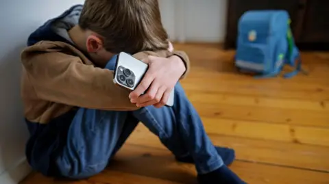 Getty Images A boy sits next to his school bag with a smartphone in his hand and hides his head. Stock photo.