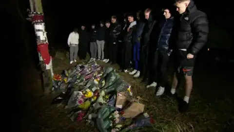 A group of lads wearing black coats and lined up looking down at floral tributes next to a post which has Grimsby football merchandise on it. It's night-time.