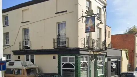 An exterior view of The Bell Inn in Cheltenham, which is on a corner and is a three-storey tall white building with a green brick frontage. Cars can be seen parked around it on the road.