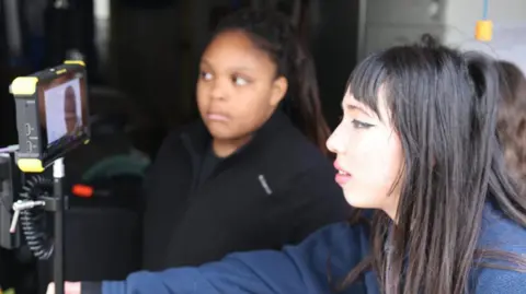 PQA Kinara and Eden watch on from behind a small screen while filming. They are both watching intently.
