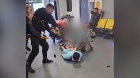 Police officer stands above man lying on floor with taser. Another officer is behind him as a woman crouches next to man on floor
