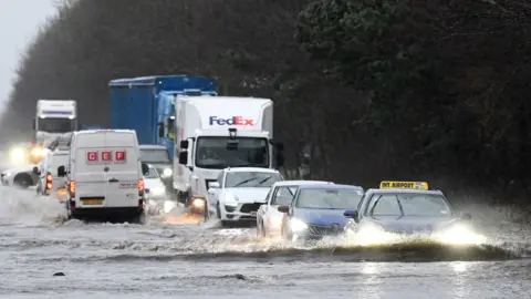Charles McQuillan/Getty Images Cars drive through heavy floods on the roads in Northern Ireland. There are several cars and lorries driving through deep water. They hae their headlights on as the light is low