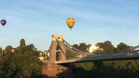 Clifton Suspension Bridge in Bristol. It is a bright sunny day with hot air balloons floating over the bridge. 