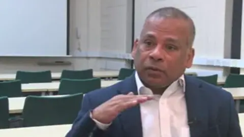 A man with a white shirt and blue blazer sits in a university lecture hall. He is surrounded by chairs and tables pointing towards a whiteboard. 