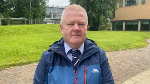 Alan Rodgers is standing outside looking into the camera. He is wearing a shirt and tie under a rain coat