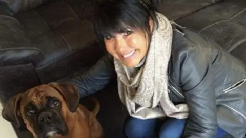 Ruth Penrose smiling with black hair, a grey scarf and black jacket and jeans, with her arm around her brown boxer dog