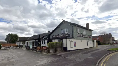 Google Maps The pub stands back from a road with a large car park on the left of the image.It has greenery decor by its entrance and a small outdoor seating area. The top part of the pub is painted pale green with a cream lower half.