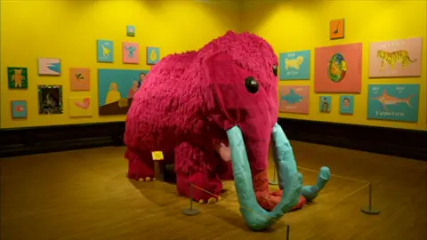  A life-size sculpture of a woolly mammoth, with pink fur, blue horns and yellow toenails in the center of the room. Colourful paintings hang on the yellow walls of an art gallery, with wooden floor.