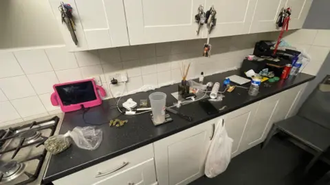 Essex Police A messy kitchen worktop, which has a knife, drugs, money and other items cluttering it. The worktop is black and the cabinets are cream.