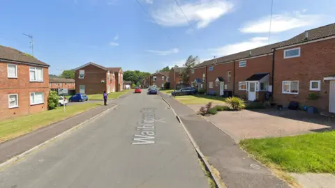 A suburban road with red brick houses on both sides and a series of grass verges.