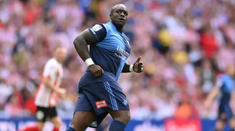 Adebayo Akinfenwa, a stocky and muscular man, jogging on a football pitch.