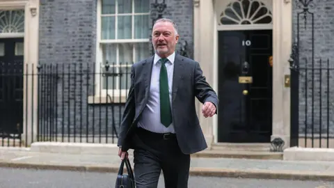 Getty Images Steve Reed in a dark suit and green tie walks outside the black front door marked number 10 on Downing Street, carrying a black bag. The image shows the street and railings in front of the government building.