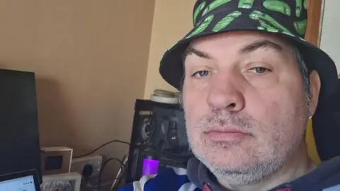 Man with a short grey beard takes a selfie wearing a black bucket hat with cartoon pickles on it. He is sat in an office environment with beige/yellow walls and a computer in front of him.