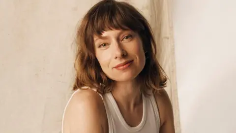 Charlotte Rutherford/SNL UK A promo shot of a woman with shoulder-length light brown hair and a fringe, wearing a white vest. She is sitting in front of a beige curtain and smiling slightly with a closed mouth.