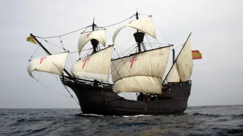 A wooden replica of a 16th Century Spanish galleon at sea with large white sails with a red cross on them 