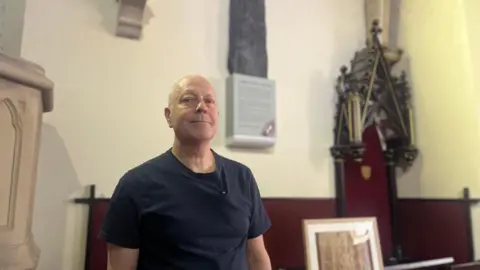 Christopher Weeks looks down at the camera, over his left shoulder mounted to the wall is a large slab of rock, known as Oruths cross. 