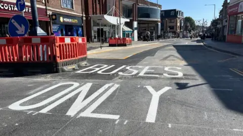 Andrew Turner/BBC New asphalt laid outside Market Gates Shopping Centre, and freshly painted "buses only" road markings in white. Orange traffic barriers are installed to the left of the image. The Market Gates shopping centre and other shop premises are visible on either side of the image.