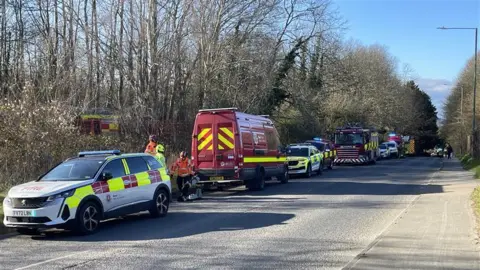 A street with emergency vehicles parked all along one side including fire engines, police cars and rescue vehicles. There are trees behind the vehicles.