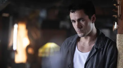 Sony Pictures Actor Jonah Hauer-King, in character, leans against a pillar inside a dimly lit restaurant with wood panelled walls. the only illumination is a lamp and shaft of sunlight behind him. He looks troubled as he stares into the middle distance.