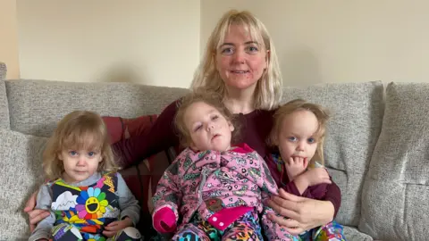Naomi pictured with shoulder length blonde hair with a fringe. She is wearing a burgundy long-sleeved top. Margot is sat on her lap wearing a colourfully printed jumper. Her other two daughters are sat on the left and right of Naomi. 