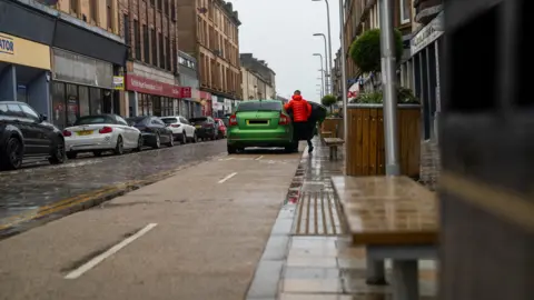 A green car parked on a two way cycle lane with a person wearing an orange jacket getting in to it.