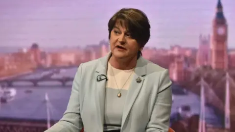 Arlene Foster, a woman with dark hair in grey suit. She is wearing a silver necklace.
