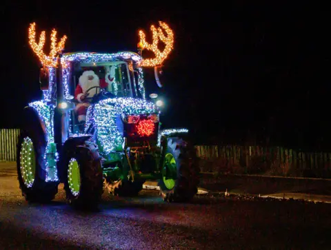 Dot Reid Tractor lit in fairy lights to make it look like Rudolph, with Santa driving and waving.