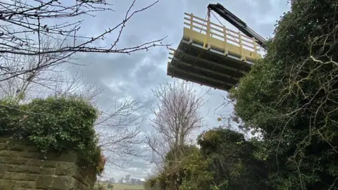 A bridge, about four metres in length, is being lowered into place from a crane. It will join two sides of an old railway