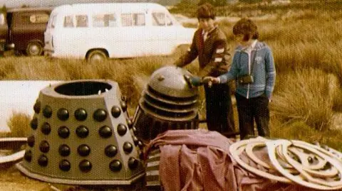 Paul and Gary Hardy-Brown Two boys stand next to the top half and base of a robotic creature (a Dalek) in a car park next to a sandy landscape