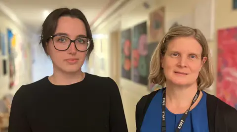 Alex Dunlop/BBC Standing in the corridor of a Sixth Form is Bethany Thomas and Kirsty Brook. On the left is Bethany Thomas who has her hair pulled back and is wearing a black long sleeved T-shirt. She is wearing glasses and is looking directly at the camera. On the right is Kirsty Brook who has blonde hair and is looking directly at the camera. She is wearing a blue T-shirt with a black cardigan over the top.