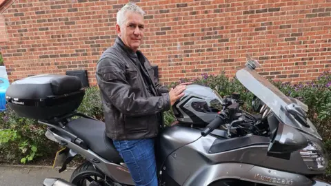 BBC/Richard Jackson A man with short grey hair sits on a Kawasaki motorbike in front of a brick wall. He is wearing a black leather jacket and blue jeans.
