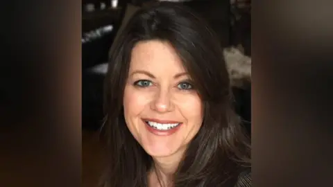 A head shot of Amanda who is looking at the camera and smiling, showing her top front teeth. She has blue eyes and dark brown shoulder lenghth brown hair. The background is blurred and dark