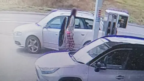 CCTV footage showing a thief wearing red and white pyjama trousers and a grey jumper filling up the tank of their silver car. Their face is blurred out. There's another car parked up next to them in the forecourt.