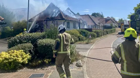 Two fire officers wearing protective pale brown overalls with hi-visibility markings in acid yellow are in a residential street lined with several bungalows. Well-maintained hedging lines the front of the properties. One of the roofs is gutted with gaping holes and billowing smoke. A fire engine with a hydraulic ladder is further up the road and a crew member is pointing a hose full of water at the burnt-out roof.  