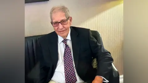 An elderly man with thinning grey hair and glasses, wearing a white pinstripe shirt, colourful tie and a dark blazer sits on a seat looking into the camera.